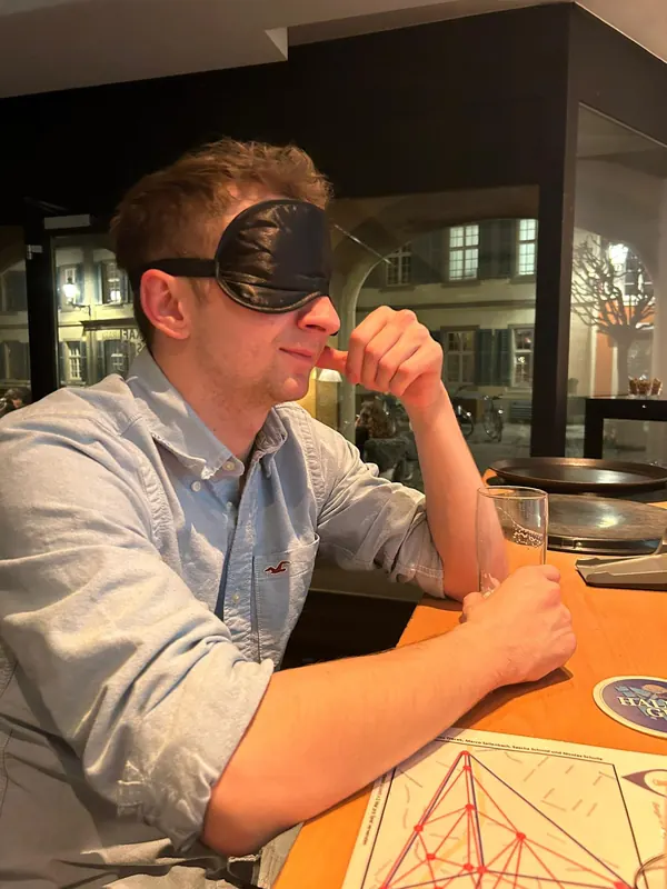 Ein Spieler mit Augenbinde sitzt an der Bar, vor ihm die gedruckte Spielkarte.