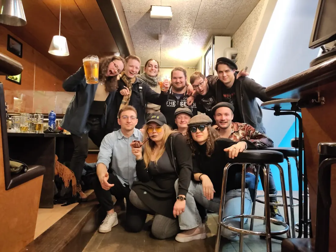 Verkleidete Spielerinnen und Spieler mit Mister-X-Mütze, Pfeife und Sonnenbrillen posieren in einer Bar.