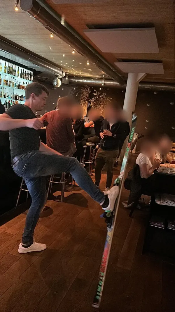 Ein Spieler kickt in einer Bar mit dem Fuss gegen einen Ski, im Hintergrund Gäste mit unkenntlich gemachten Gesichtern.